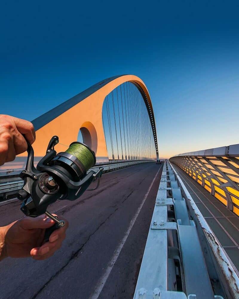 Person holding a fishing reel on a modern bridge at sunset.