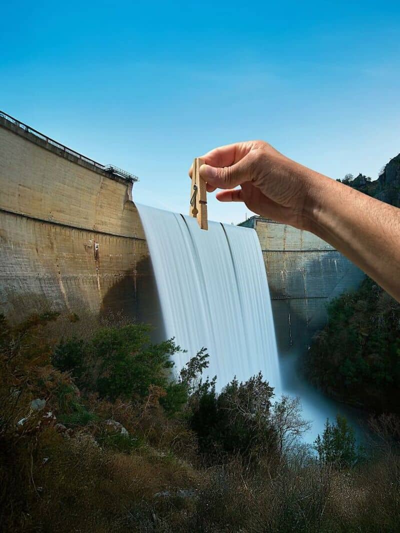 Hand holding a clothespin near a dam with water flowing over it.