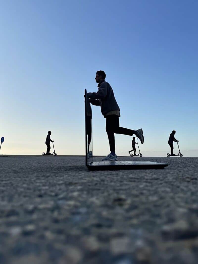 Man on a laptop with people riding scooters in the background under a clear sky.