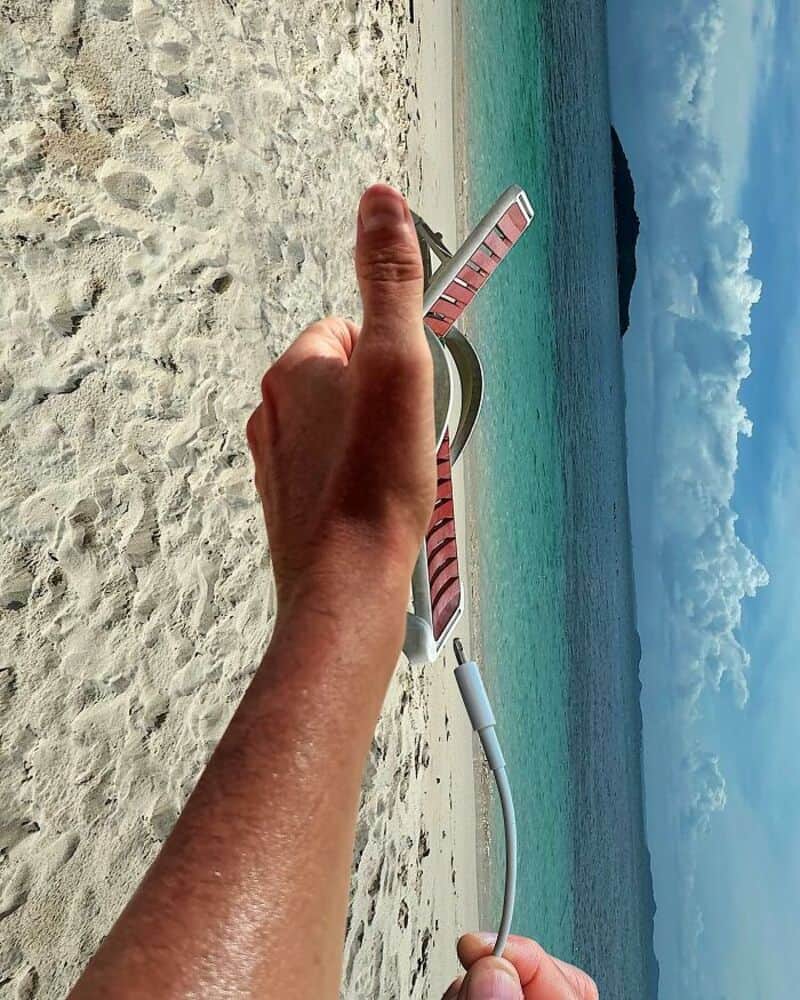 Person holding a pink and white striped flip-flop on a sandy beach with ocean and sky in background.