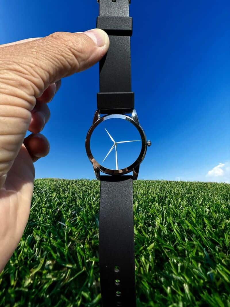 Hand holding a black wristwatch with a wind turbine design on the face against a blue sky.