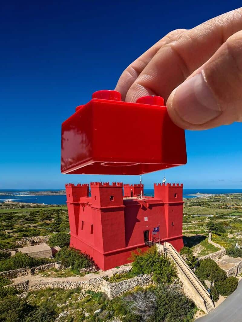 Giant red LEGO brick held above a red castle by a hand against a blue sky.