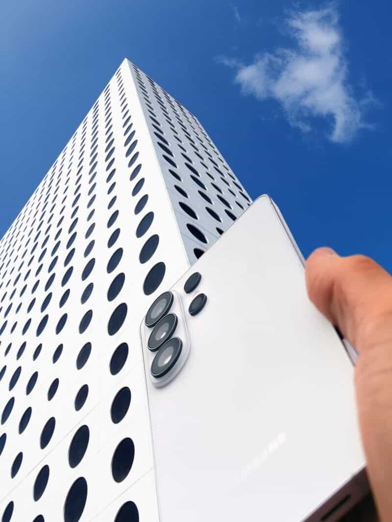 A hand holding a smartphone in front of a tall, white building with circular windows.