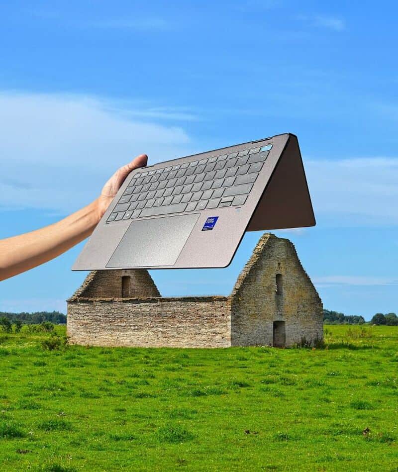 Person holding a laptop over a small stone house in a field under a blue sky.
