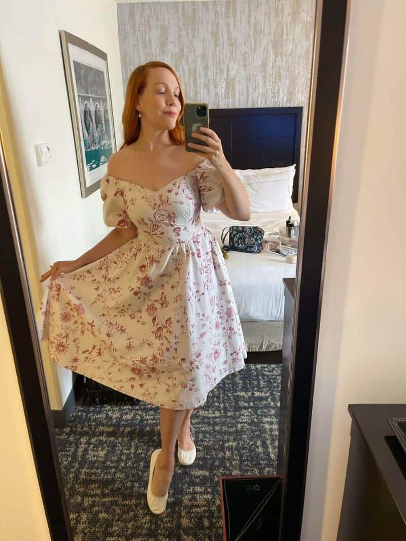 Woman in floral dress taking mirror selfie in hotel room.