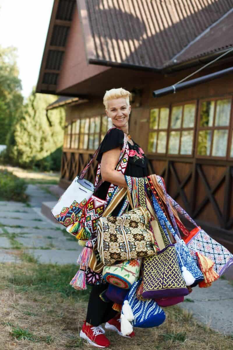 Smiling woman with short blonde hair carrying many colorful upcycled bags outside.