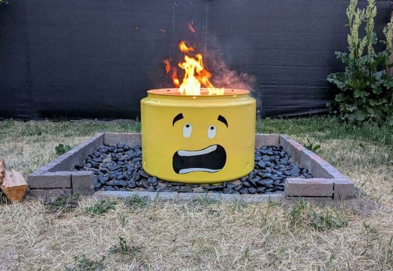 Yellow barrel with a surprised face, on fire, in a garden.