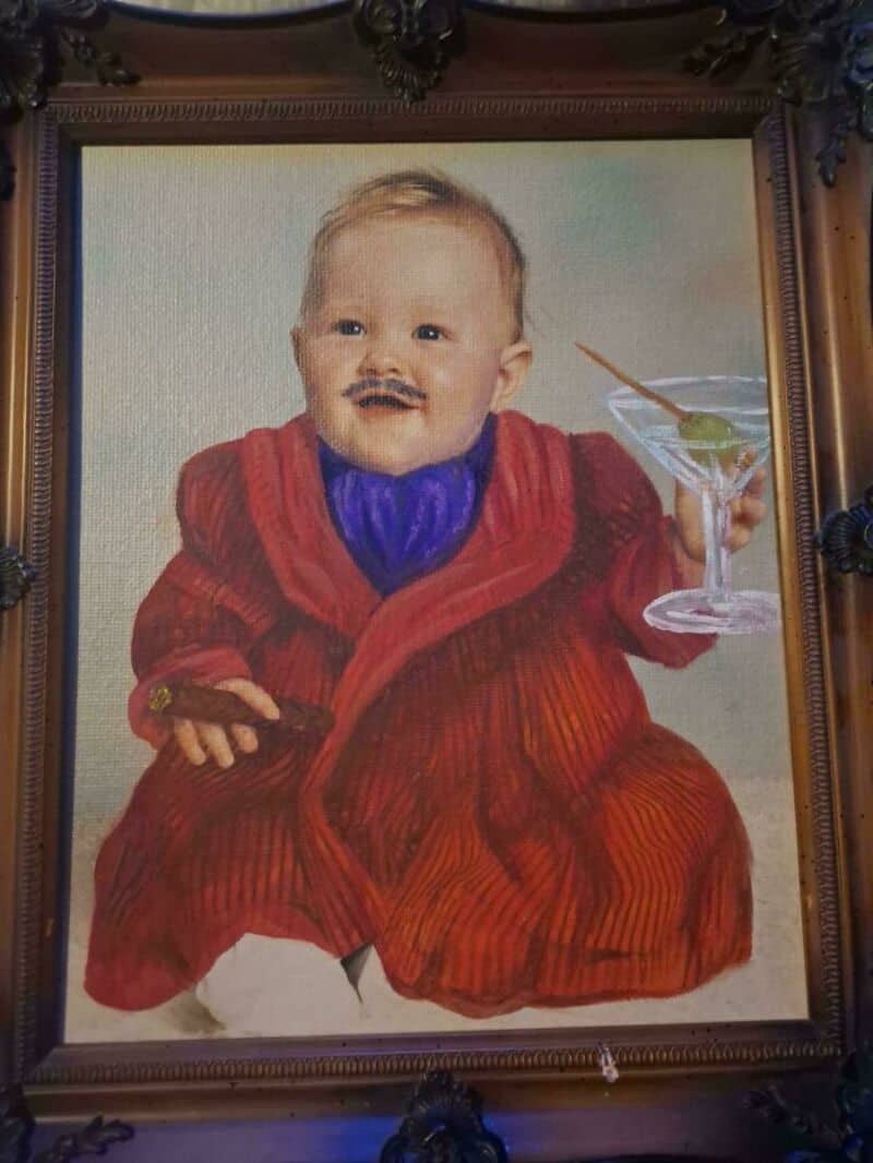 Painting of a baby in a red robe holding a cigar and a cocktail glass.