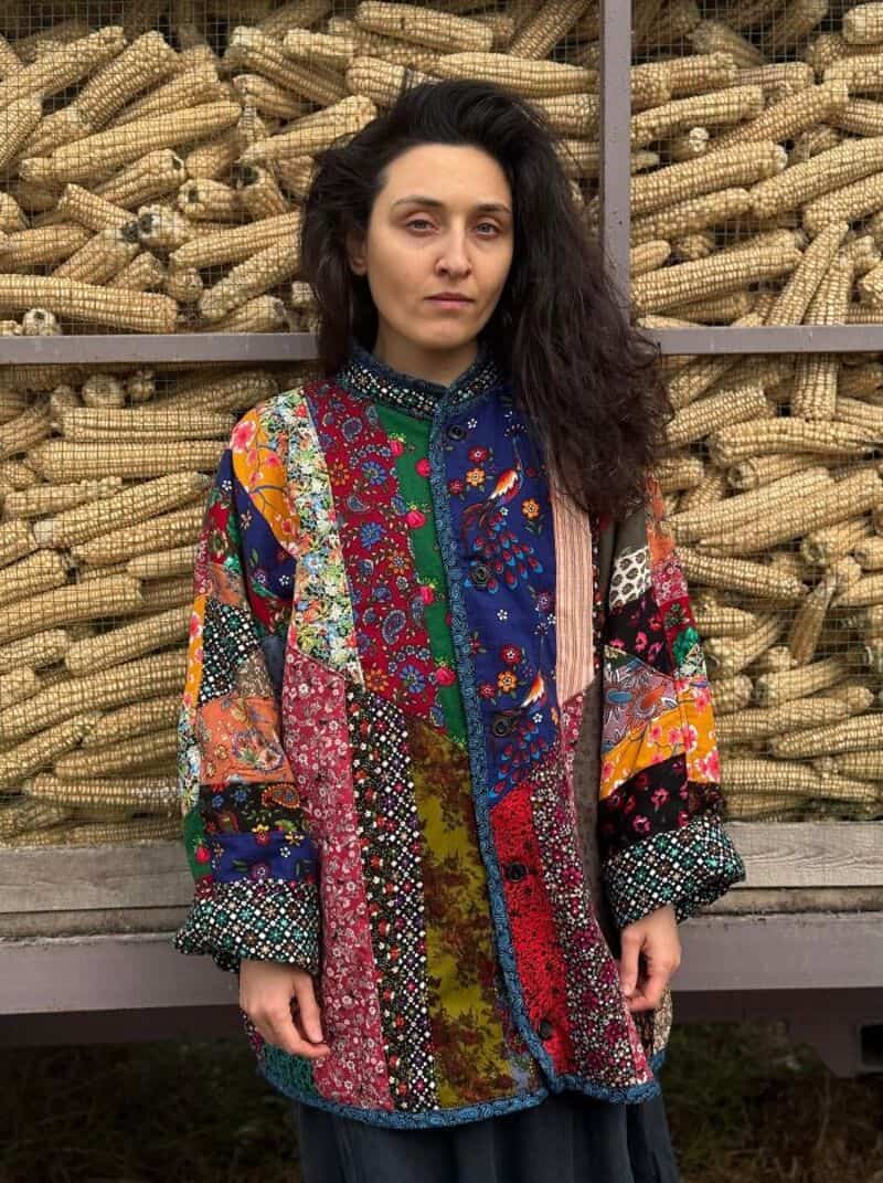 Woman with dark curly hair wearing a colorful patchwork coat in front of a corn display.
