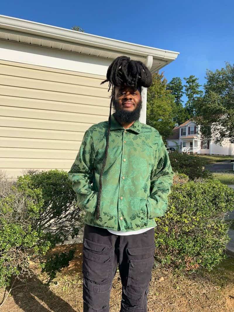 Young man with dreadlocks in a green patterned jacket outside.
