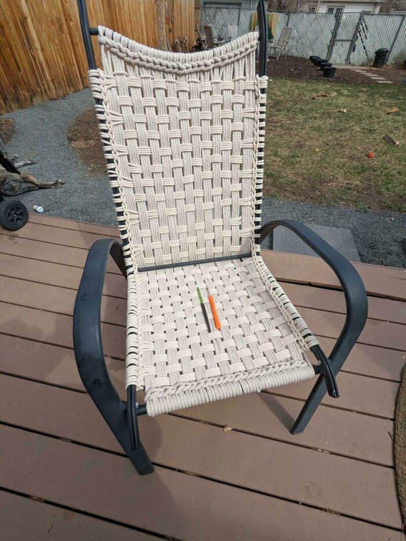 Woven outdoor chair with black metal frame on wooden deck, backyard in background.