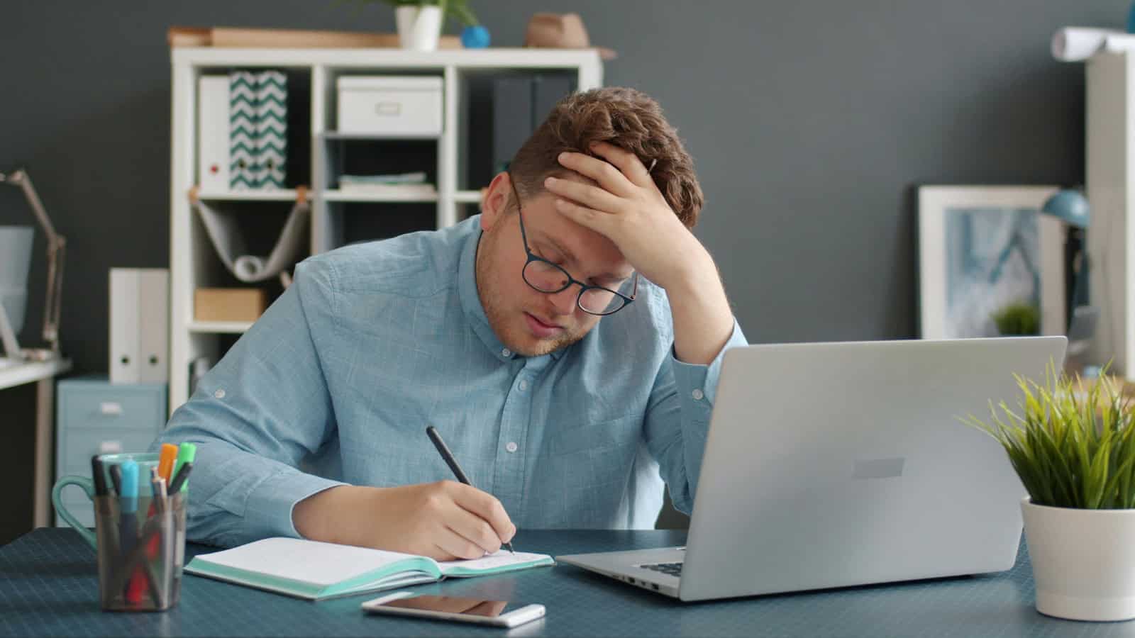 Frustrated man working at desk with laptop and notebook in office.