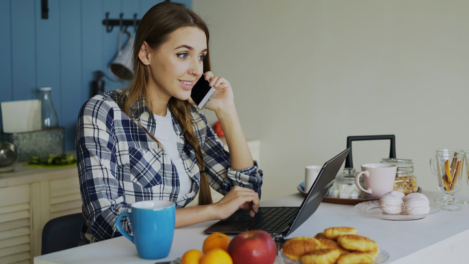 Young woman on phone at kitchen table with laptop and snacks.