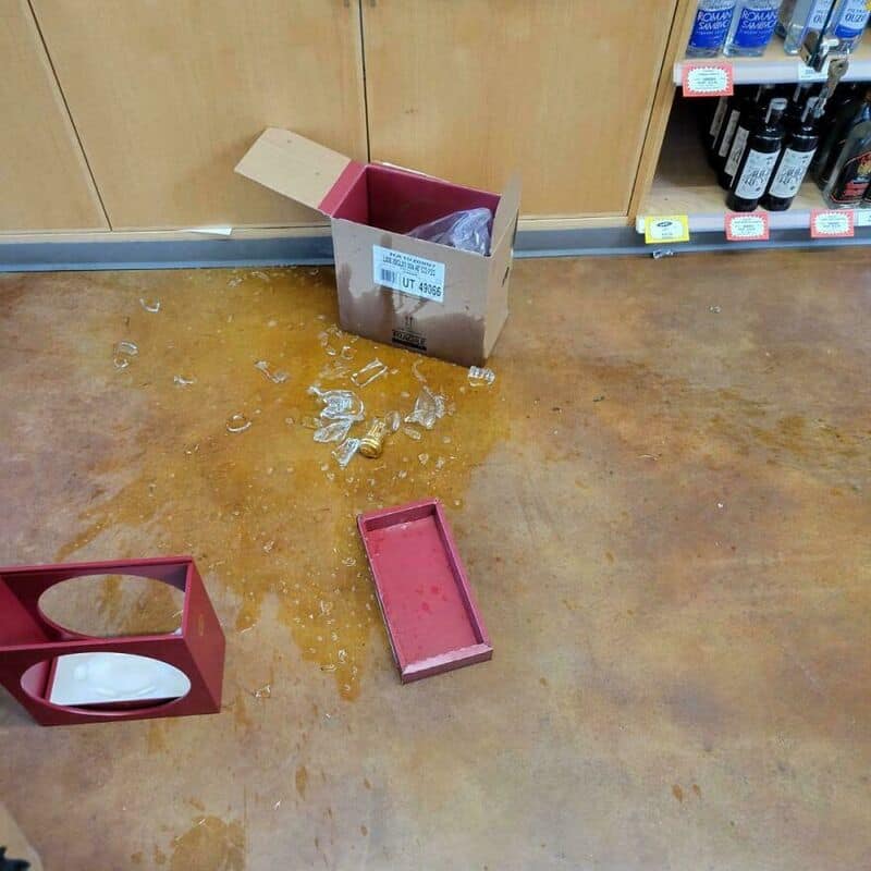 Spilled liquid and broken glass on store floor near an open box and empty tray.