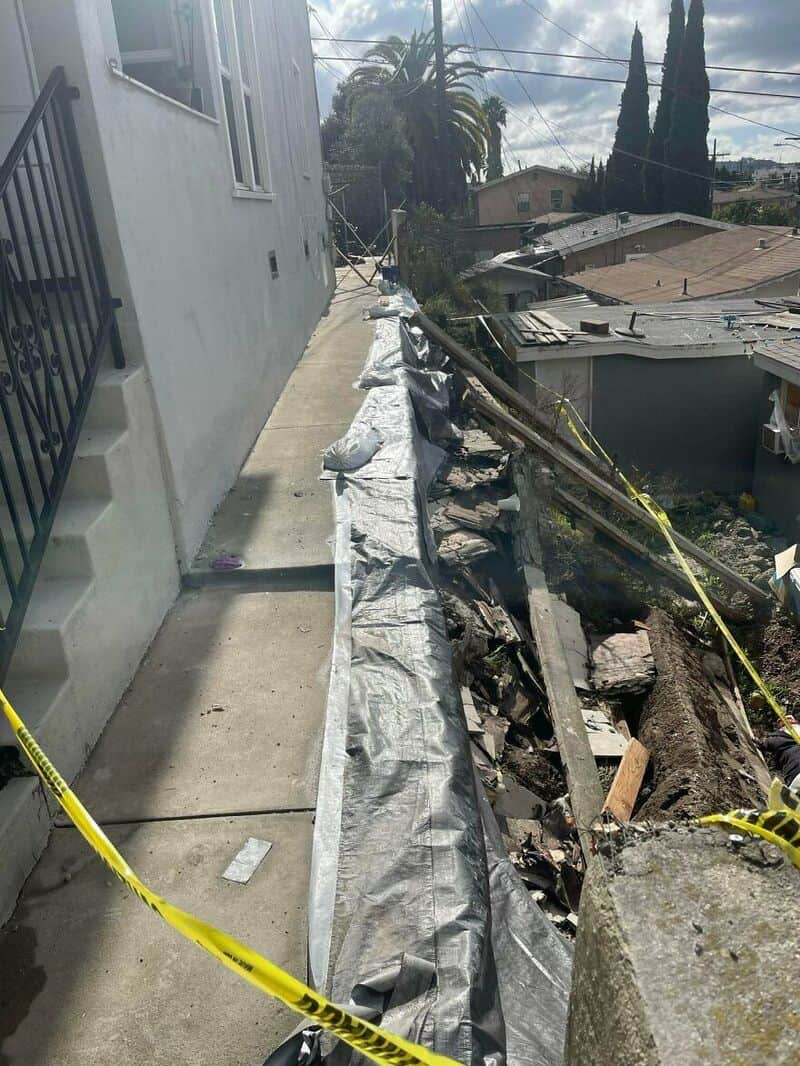Damaged sidewalk and collapsed retaining wall with debris and caution tape.