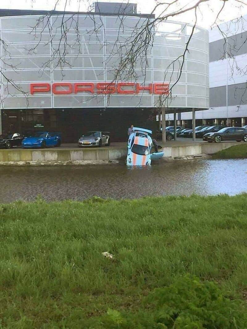 Car crashed into water in front of Porsche dealership, with cars parked inside.