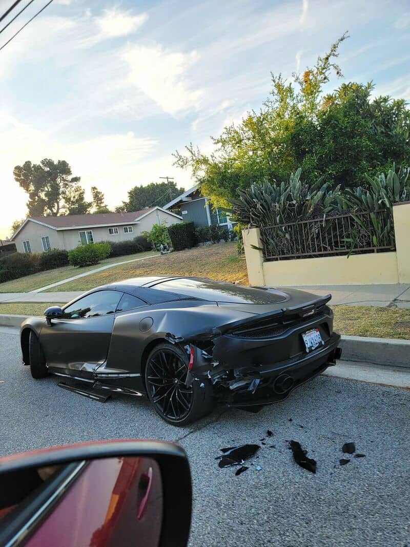 Black sports car with rear damage on the street, debris nearby, residential area in background.