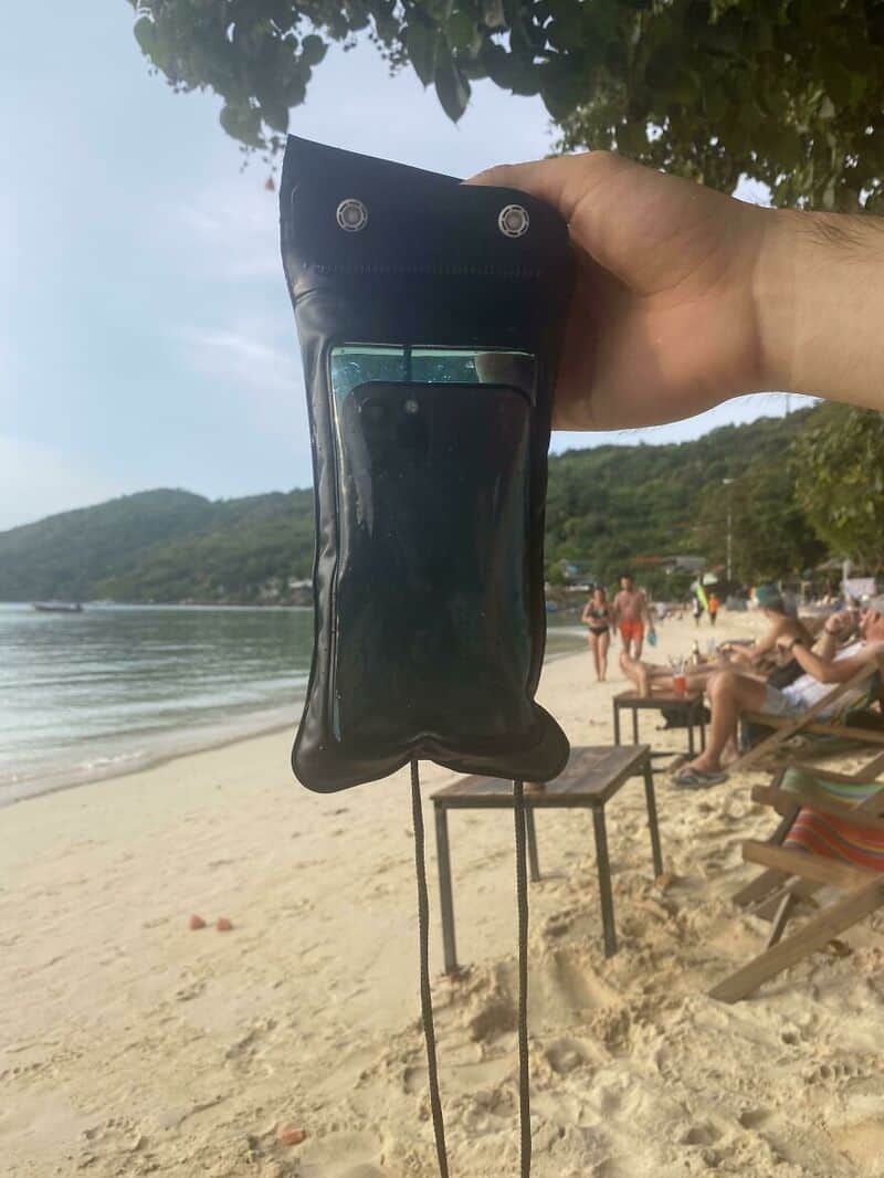 A black waterproof pouch with a transparent window, hanging from a person's hand at the beach.