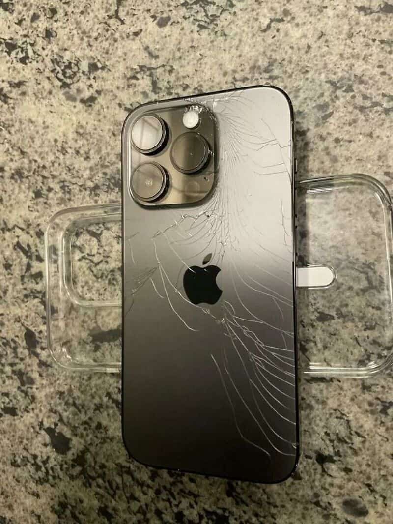Damaged iPhone with cracked back glass on a speckled surface.