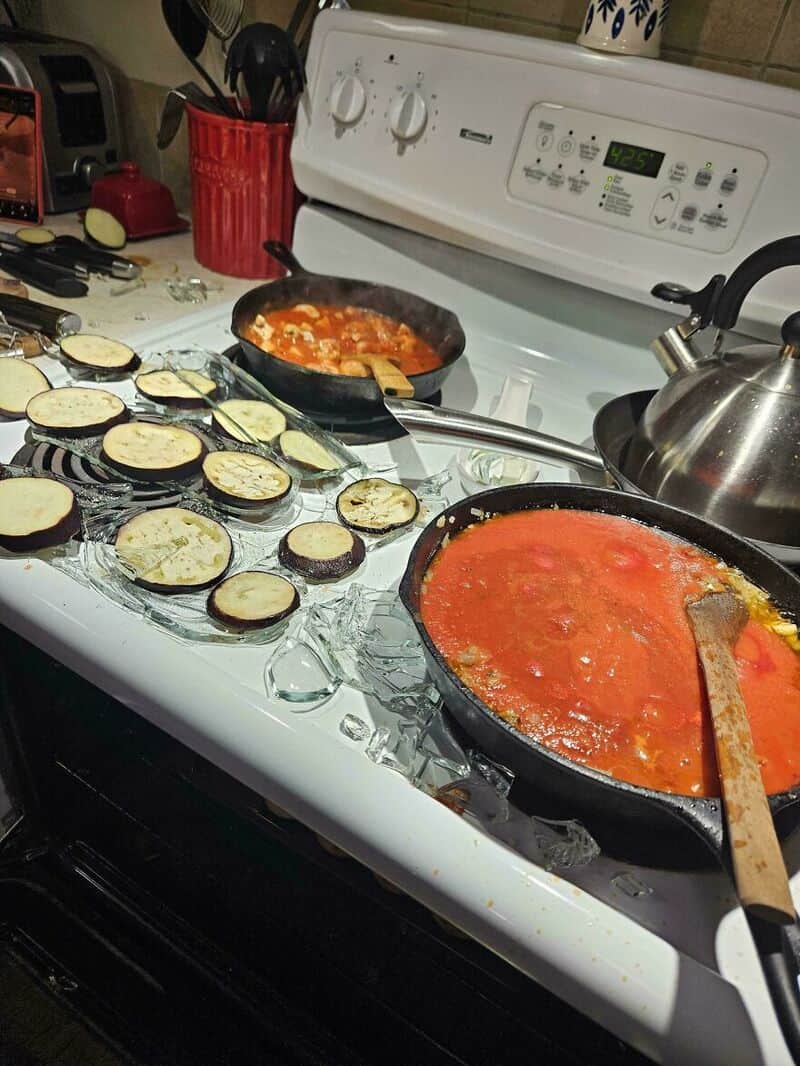 Eggplant slices on a baking sheet and two pans of tomato sauce on stove.