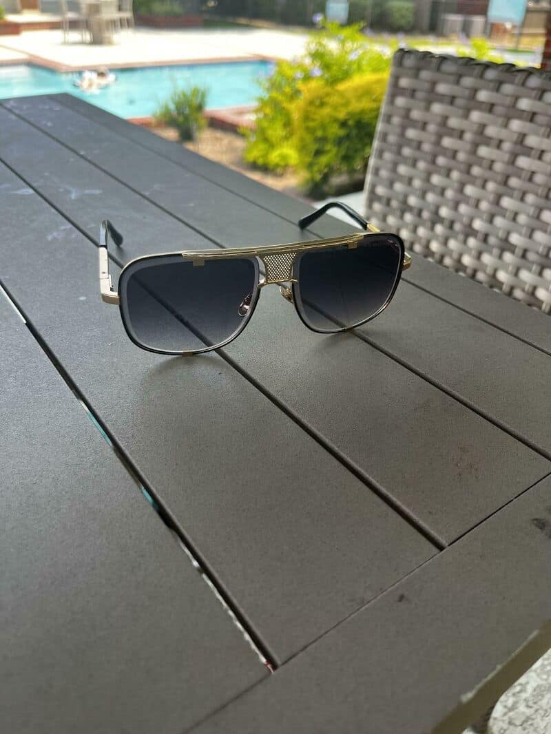 Sunglasses on a dark table near a pool with greenery in the background.