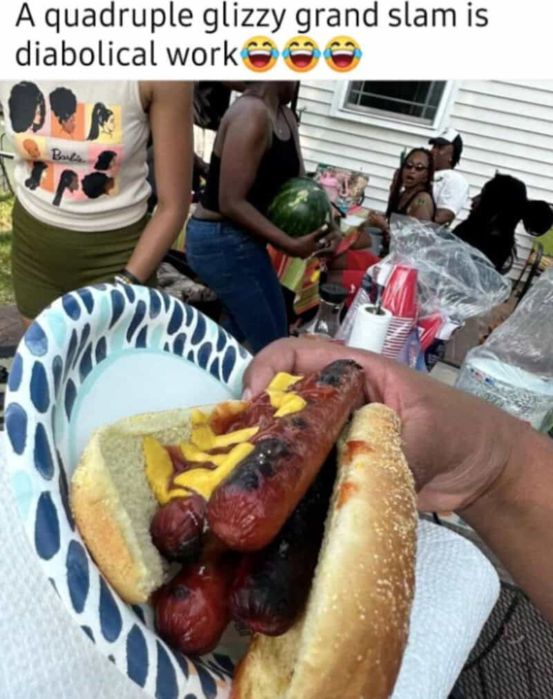 A hot dog with multiple sausages and mustard, held in front of a group at a barbecue.