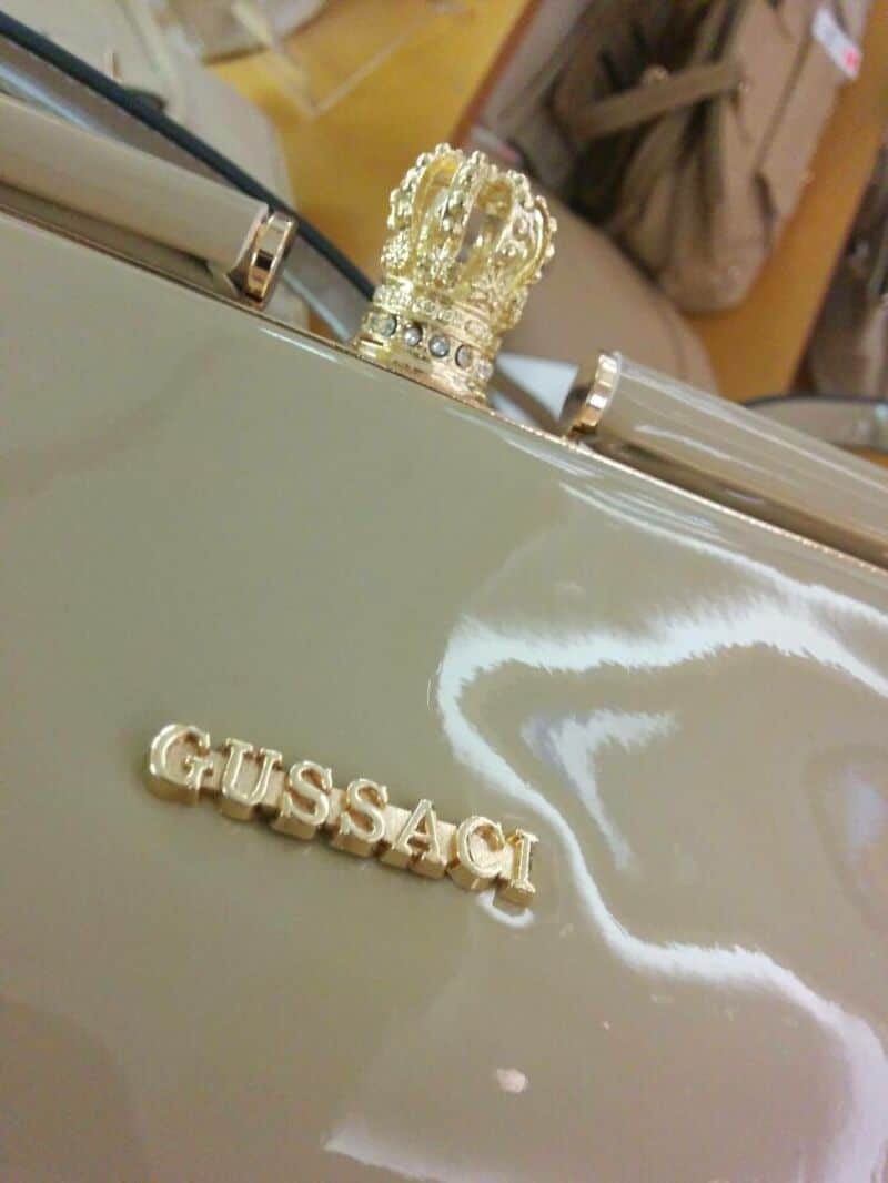 Close-up of a beige handbag with a gold 'GUSSACI' logo and a crown ornament.