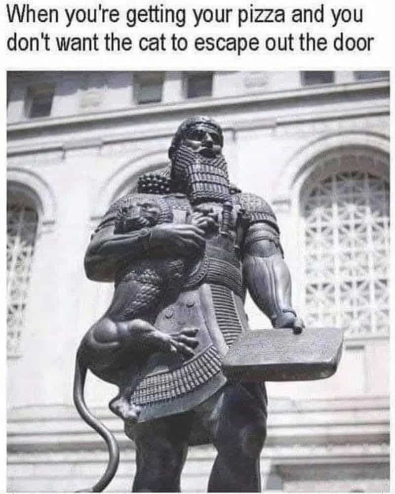Statue of a warrior with a cat on its arm, holding a pizza box, in front of a building.