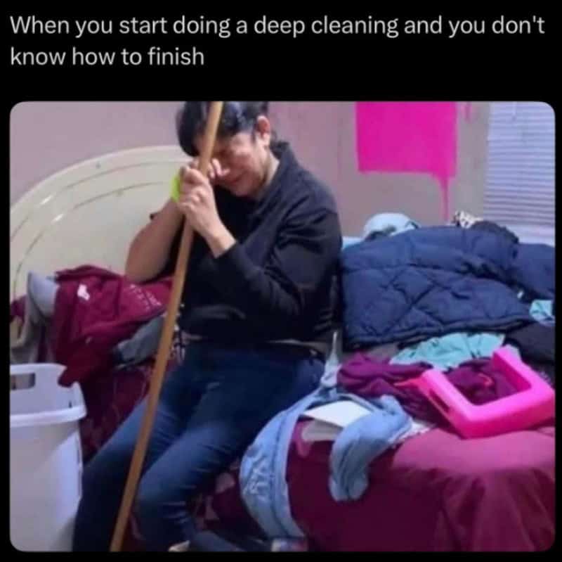 Person cleaning with a broom, surrounded by clothes and laundry on bed and floor.
