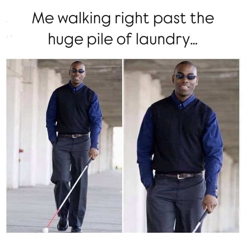 Man walking past laundry pile with a cane, wearing sunglasses, blue shirt, and black vest.