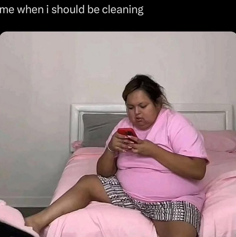 Woman in pink shirt sitting on bed, looking at her phone, with text 'me when i should be cleaning'.