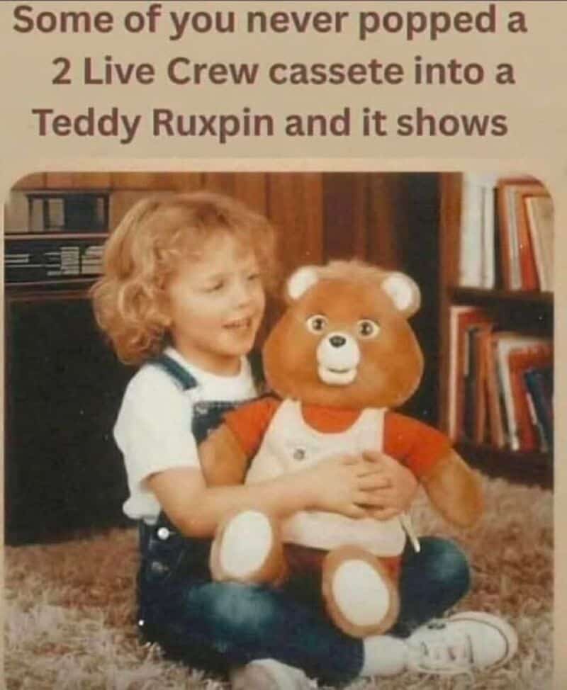 Child with curly hair holding a Teddy Ruxpin plush toy in a room with bookshelves.