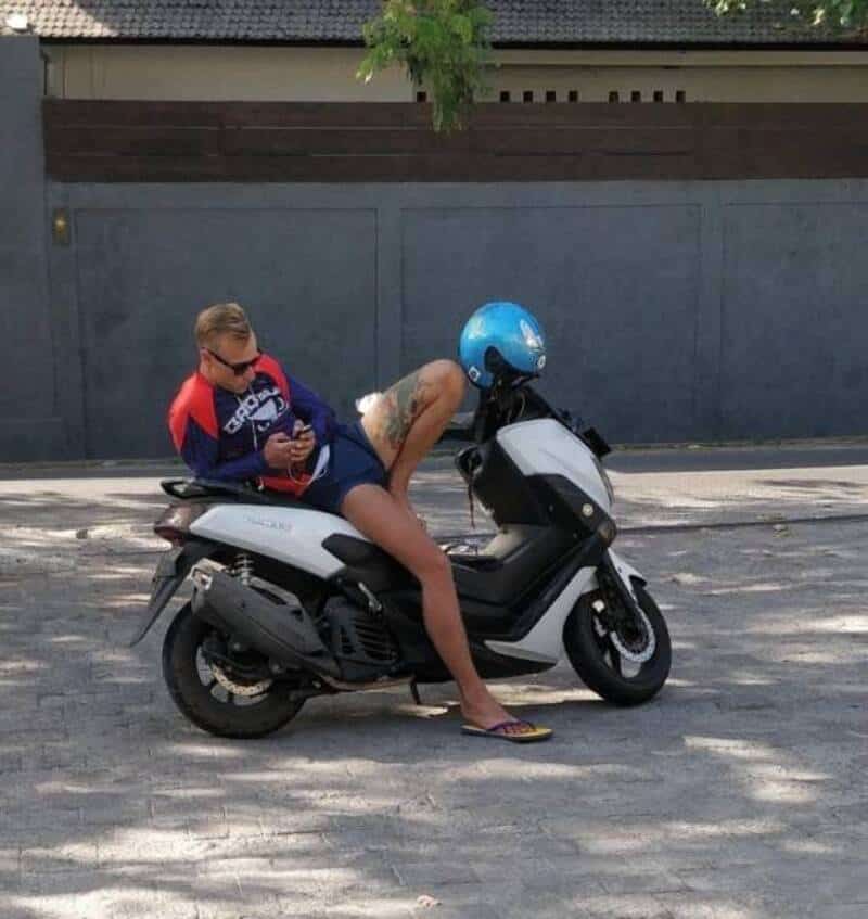 Person lying on scooter with helmet, using phone, in front of a wall.