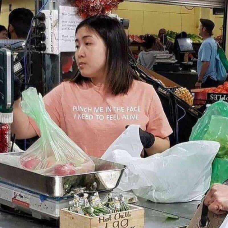 Young woman at a market stall with a shirt that says 'PUNCH ME IN THE FACE I NEED TO FEEL ALIVE'.