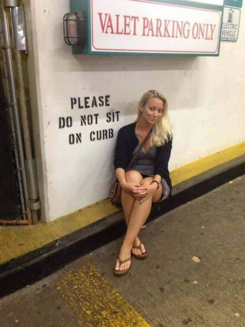 Woman sitting on curb near valet parking sign, ignoring 'Do not sit on curb' sign.