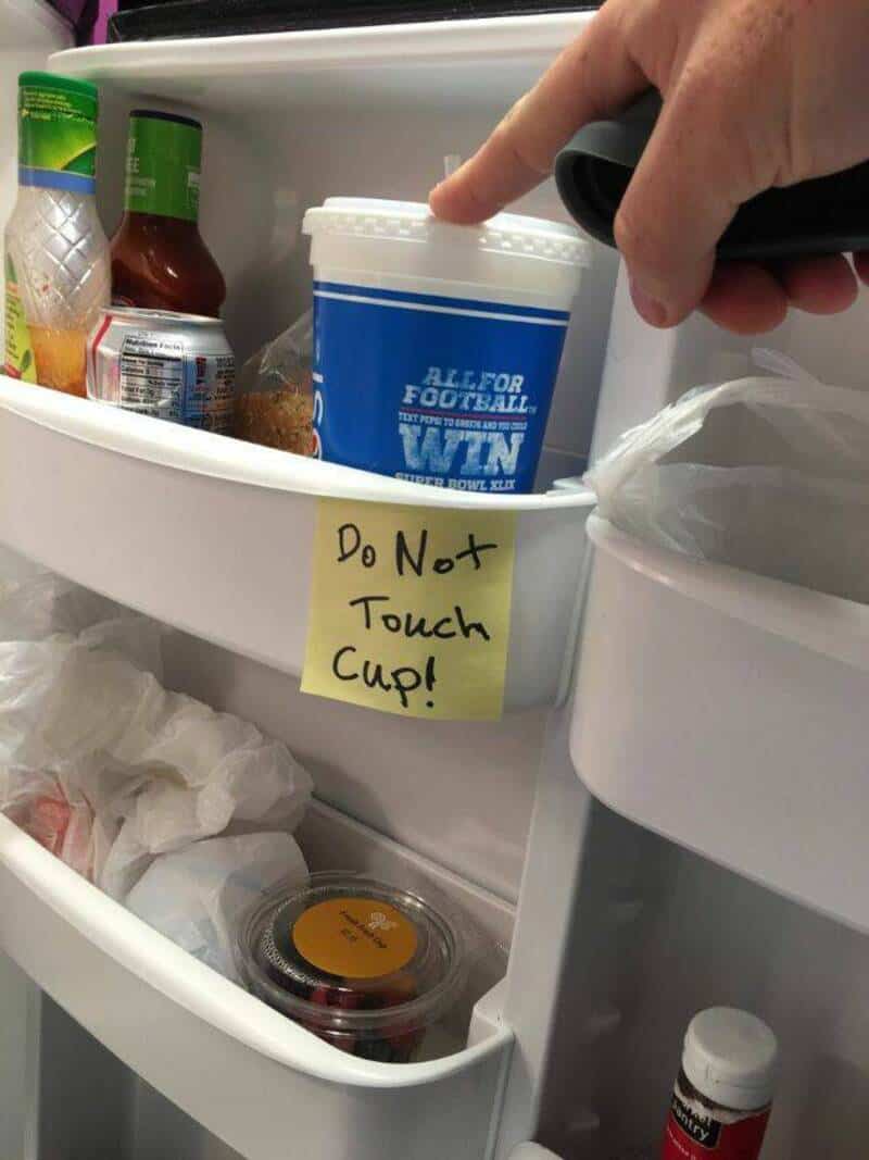 Hand reaching into fridge, touching a cup with a note that says 'Do Not Touch Cup!'.