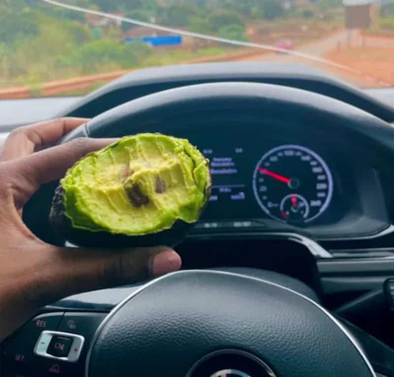 Person holding a half avocado in front of a car dashboard on a dirt road.