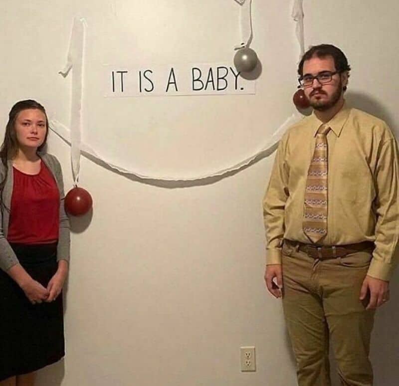 Two people stand in front of a wall with a sign that says 'IT IS A BABY.'.
