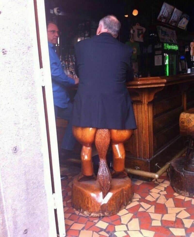 Man in suit sitting on a wooden sculpture of a humorous animal figure at a bar.