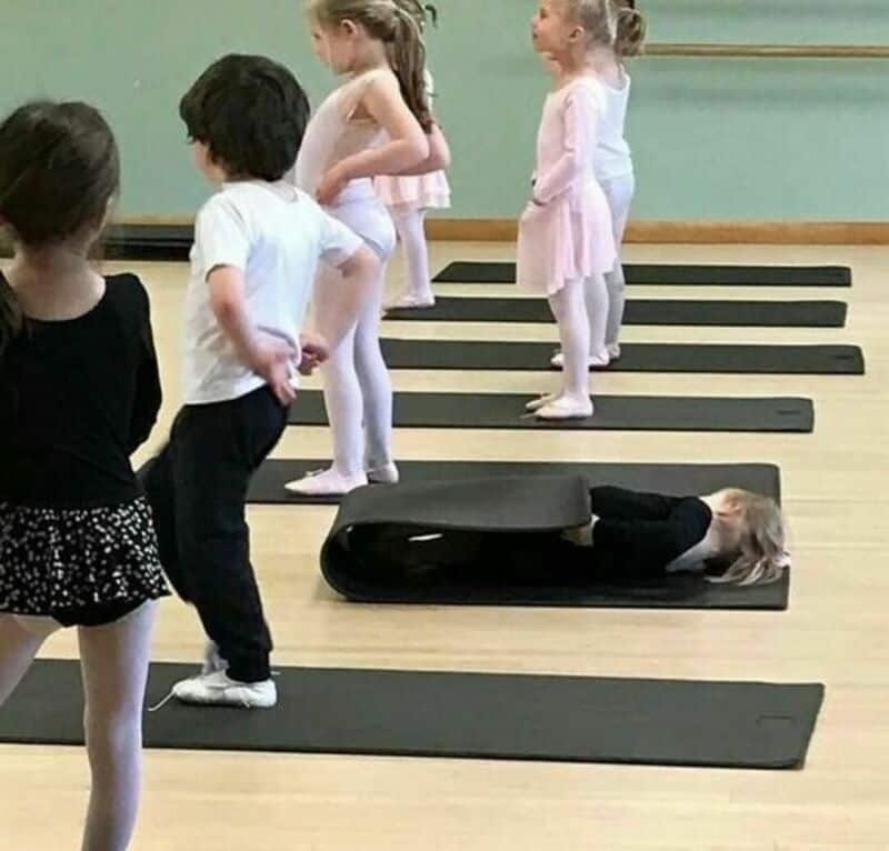 Children in a dance class with one lying on a mat, others standing in line.