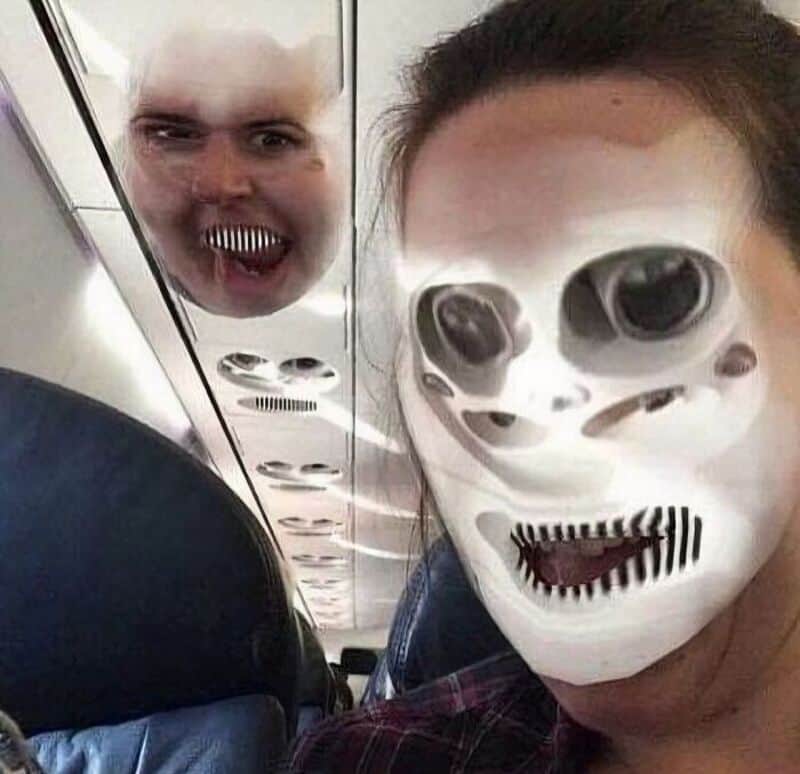 Person wearing a skull mask with a reflection of a face with sharp teeth in the airplane window.