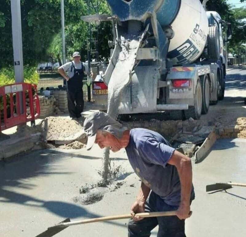 Man shoveling concrete as a cement mixer pours it on the street.