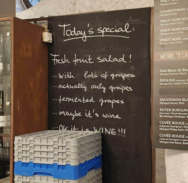 Chalkboard menu with humorous wine joke and stacked crates in front.