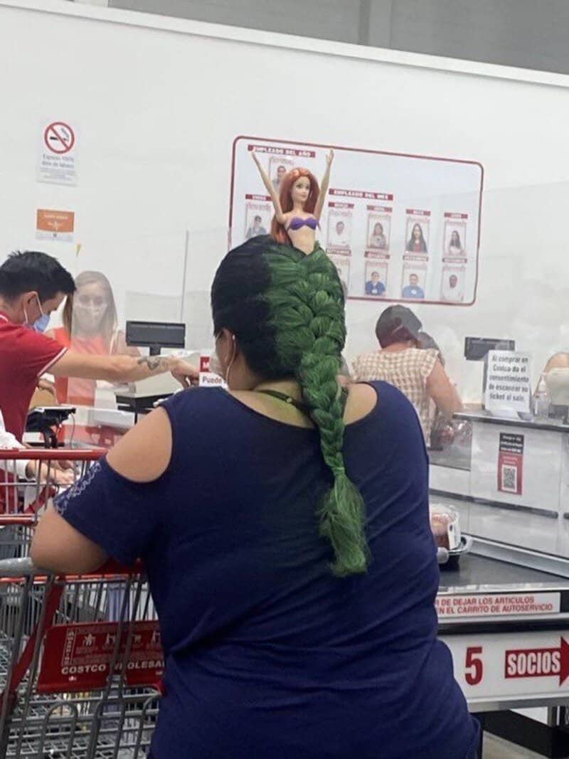 Woman with green braided hair at checkout in store, with a doll on her head.