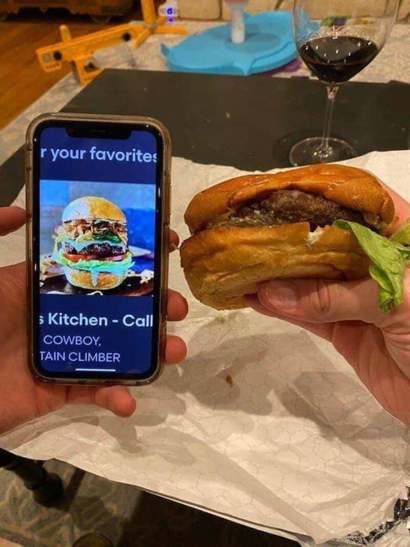 Person holding a phone showing a burger image next to a real burger and a glass of wine.