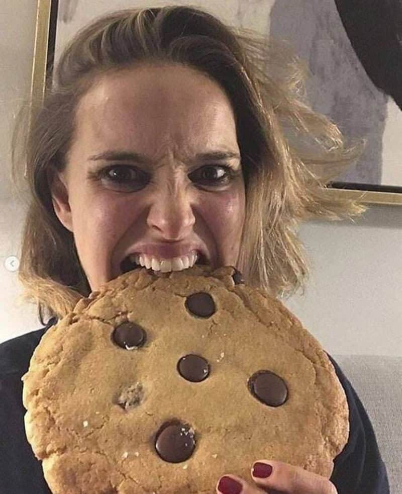 Woman with messy hair biting into a large chocolate chip cookie.