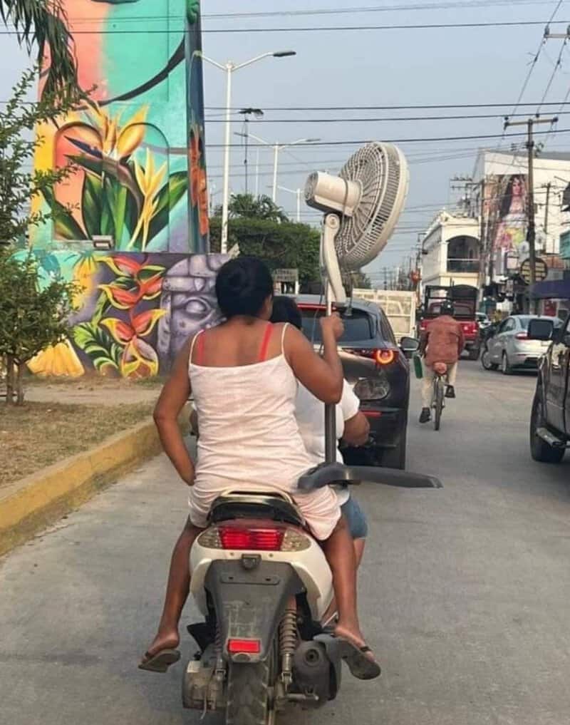Woman riding motorcycle with a large fan attached to her hand on a busy street.