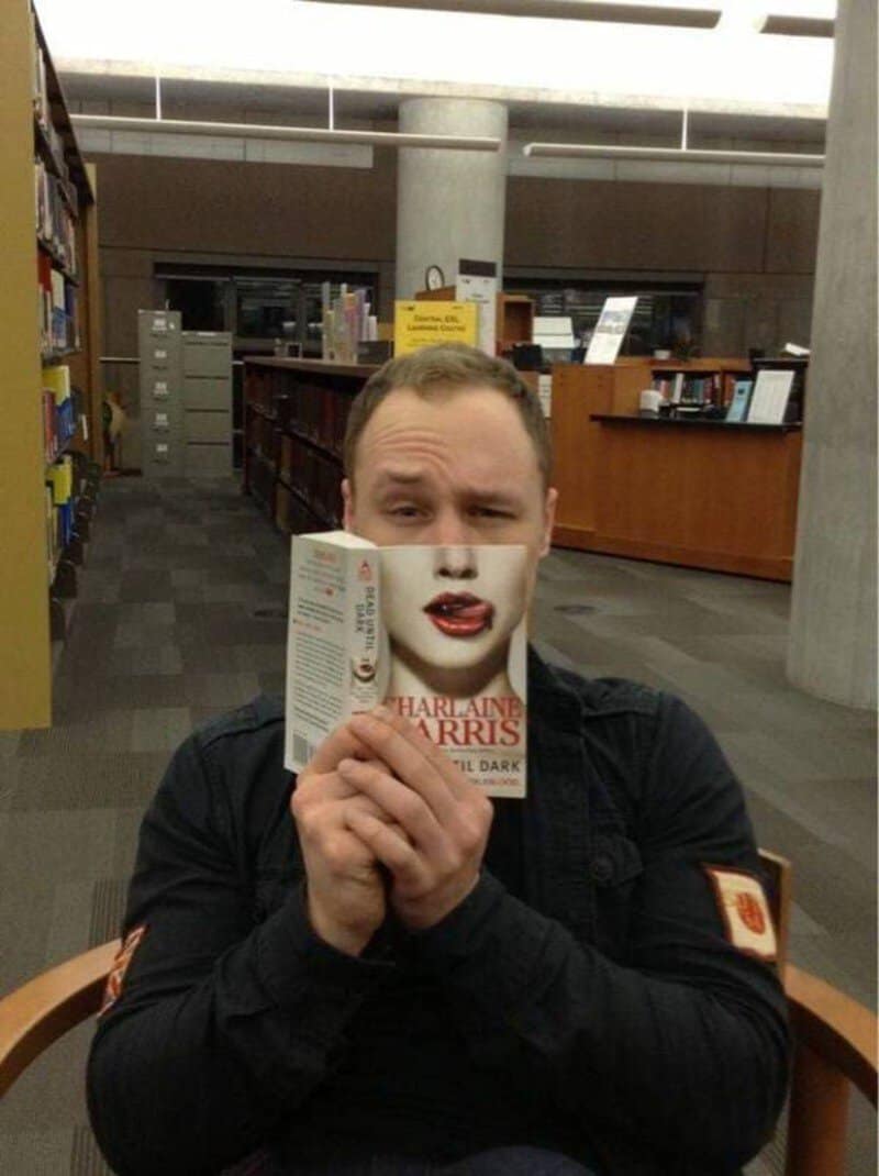 Person with makeup holding a book in a library, with a serious expression.