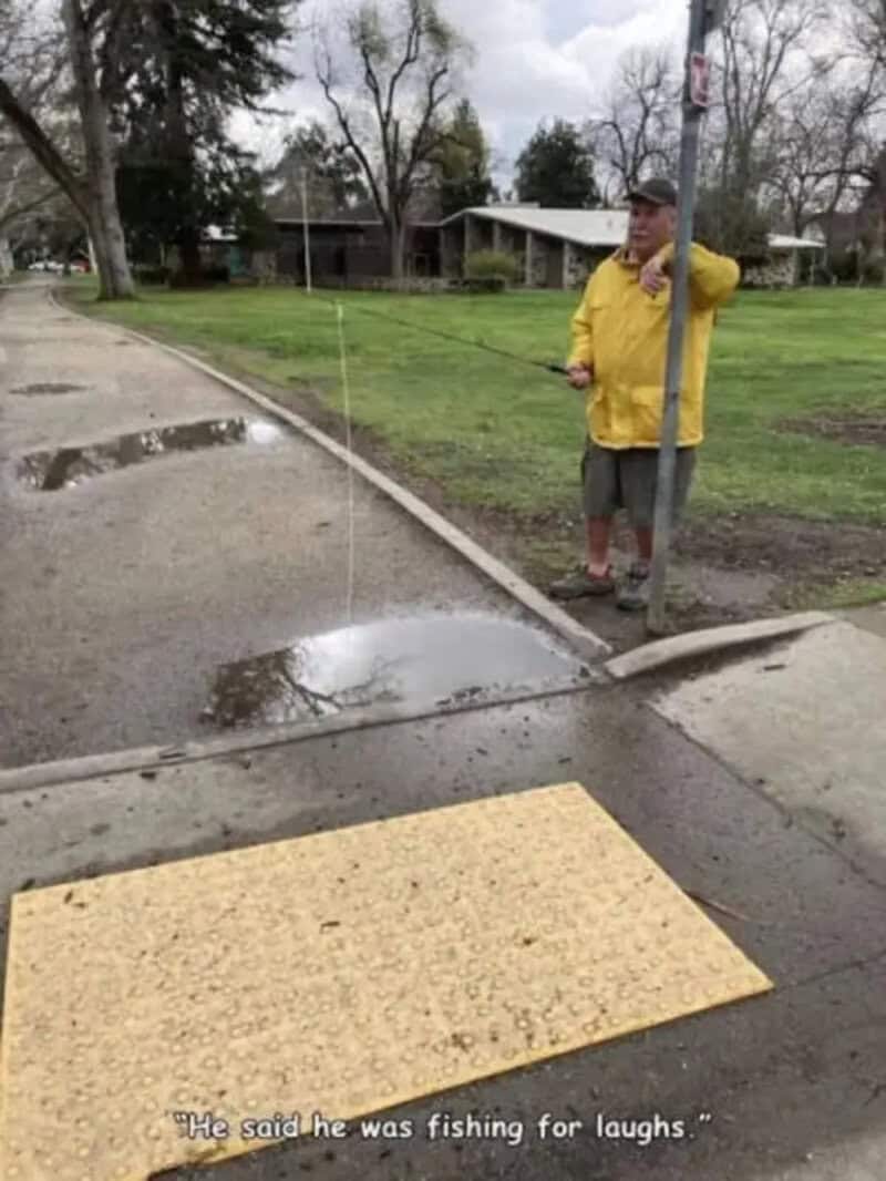 Man in yellow jacket fishing for laughs at a puddle on the sidewalk.