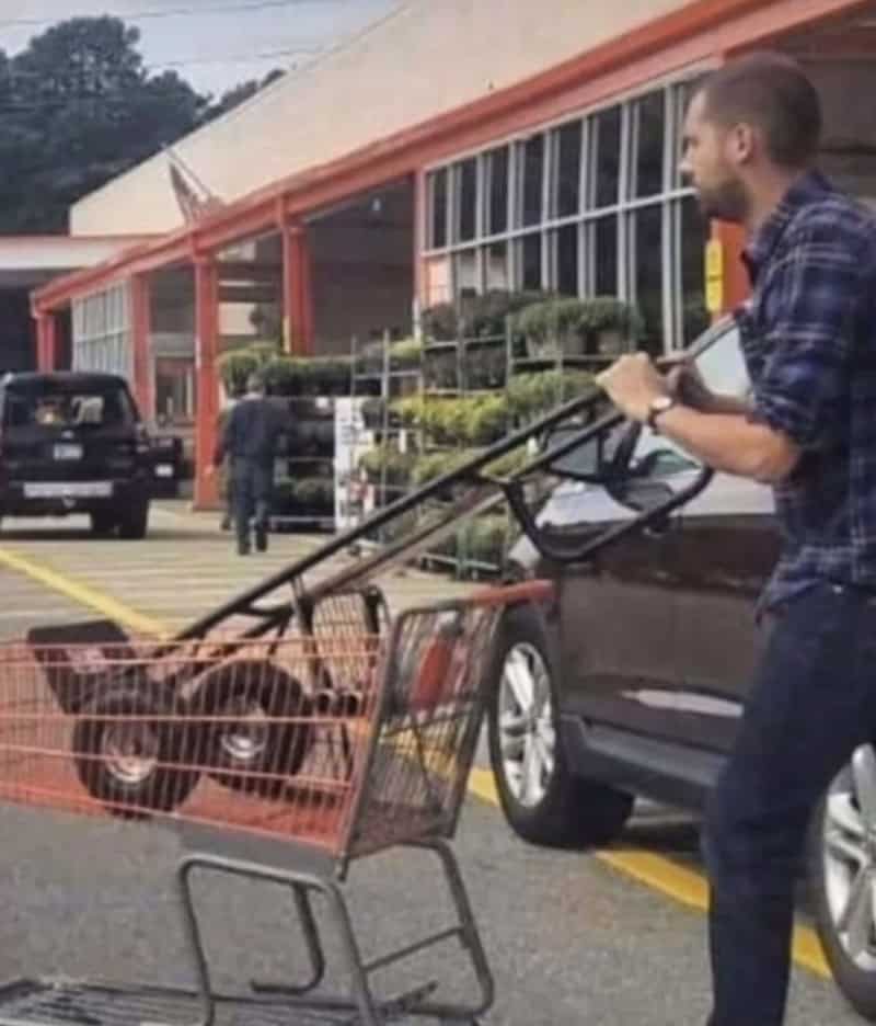 Man pushing shopping cart with a dog inside it in a parking lot.